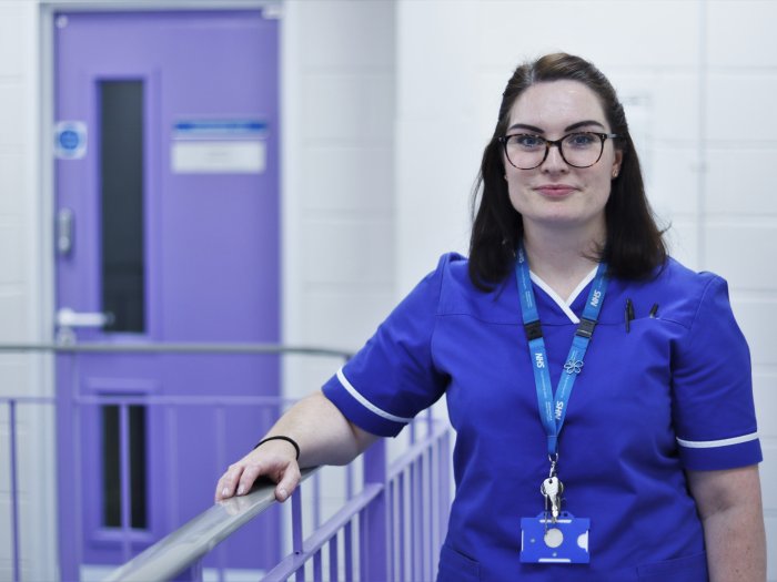 Woman in scrubs at top of staircase, hand resting on bannister, looking at camera