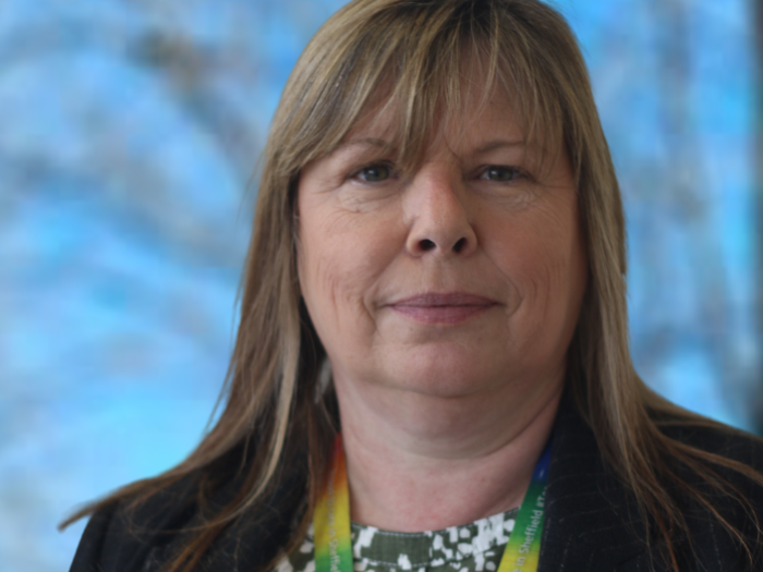 Salli Midgley in a suit jacket with rainbow lanyard around her neck against a blue sky with trees