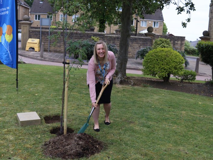 Sarah Ellison smiling while holding a spade to a pile of soil next to a hole in the ground in which a tree has been planted