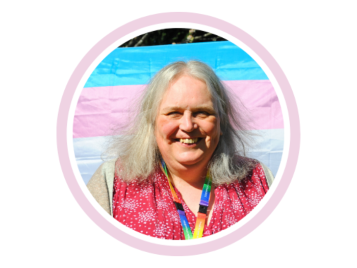 Helen Goodson, a peer support worker working in the Gender Identity Clinic, smiles whilst standing in front of a trans flag (which is made up of a blue stripe, then a pink stripe, then a white stripe, then a pink stripe, and then a blue stripe)