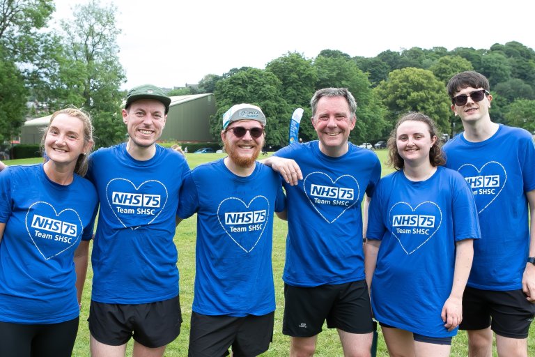 Six people stand in a park arm in arm in matching NHS75 heart t-shirts and running gear
